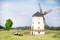 Un modèle d'un moulin aux ailes tournantes se trouve dans un champ. Au premier plan, un petit tracteur avec une remorque est visible.