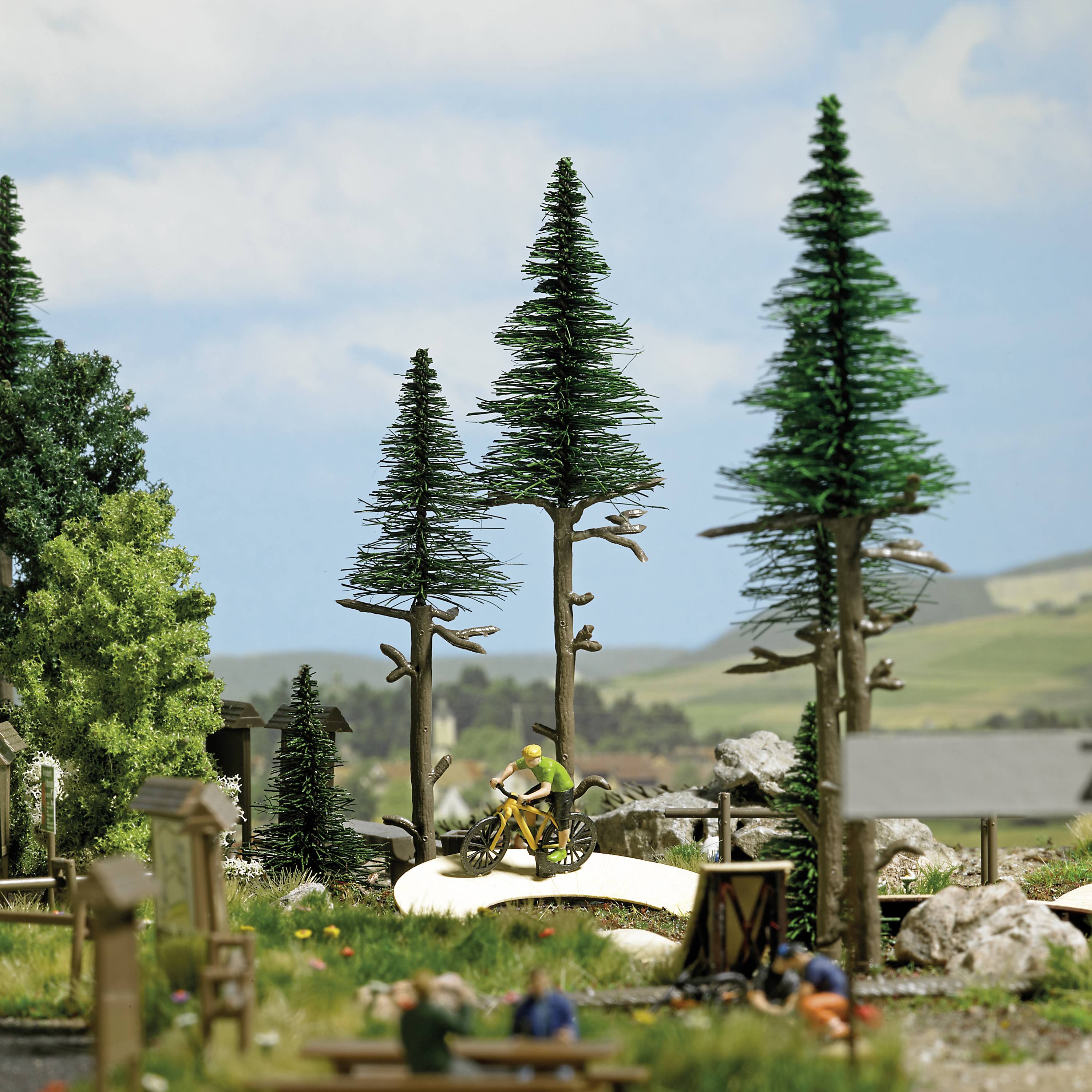 Un paysage miniature montre un petit parc avec de grands arbres et deux cyclistes sur un chemin sinueux. Des collines sont visibles en arrière-plan.