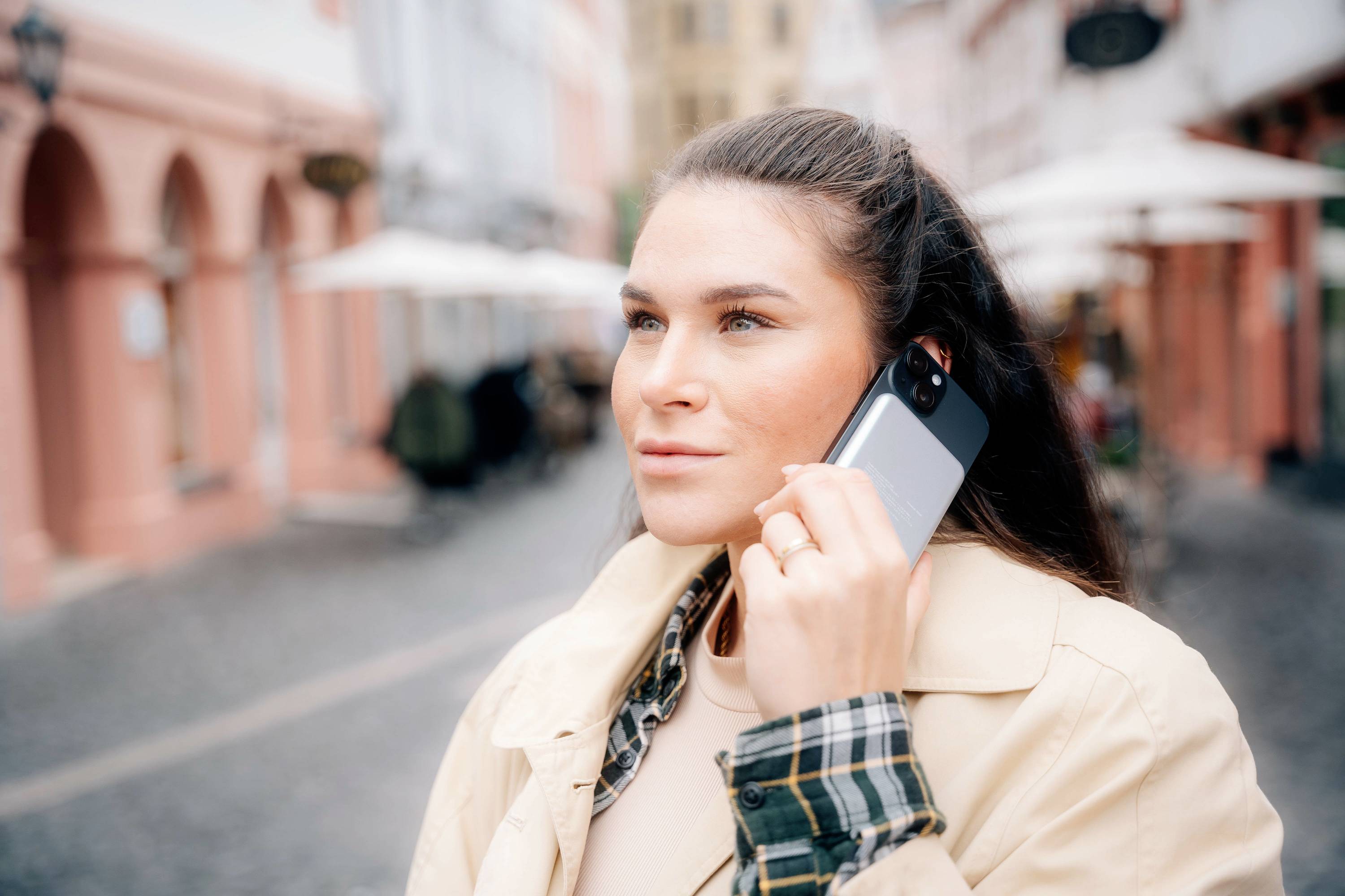 Une personne dans un environnement urbain tient un smartphone contre son oreille et semble téléphoner. Des bâtiments sont visibles en arrière-plan.