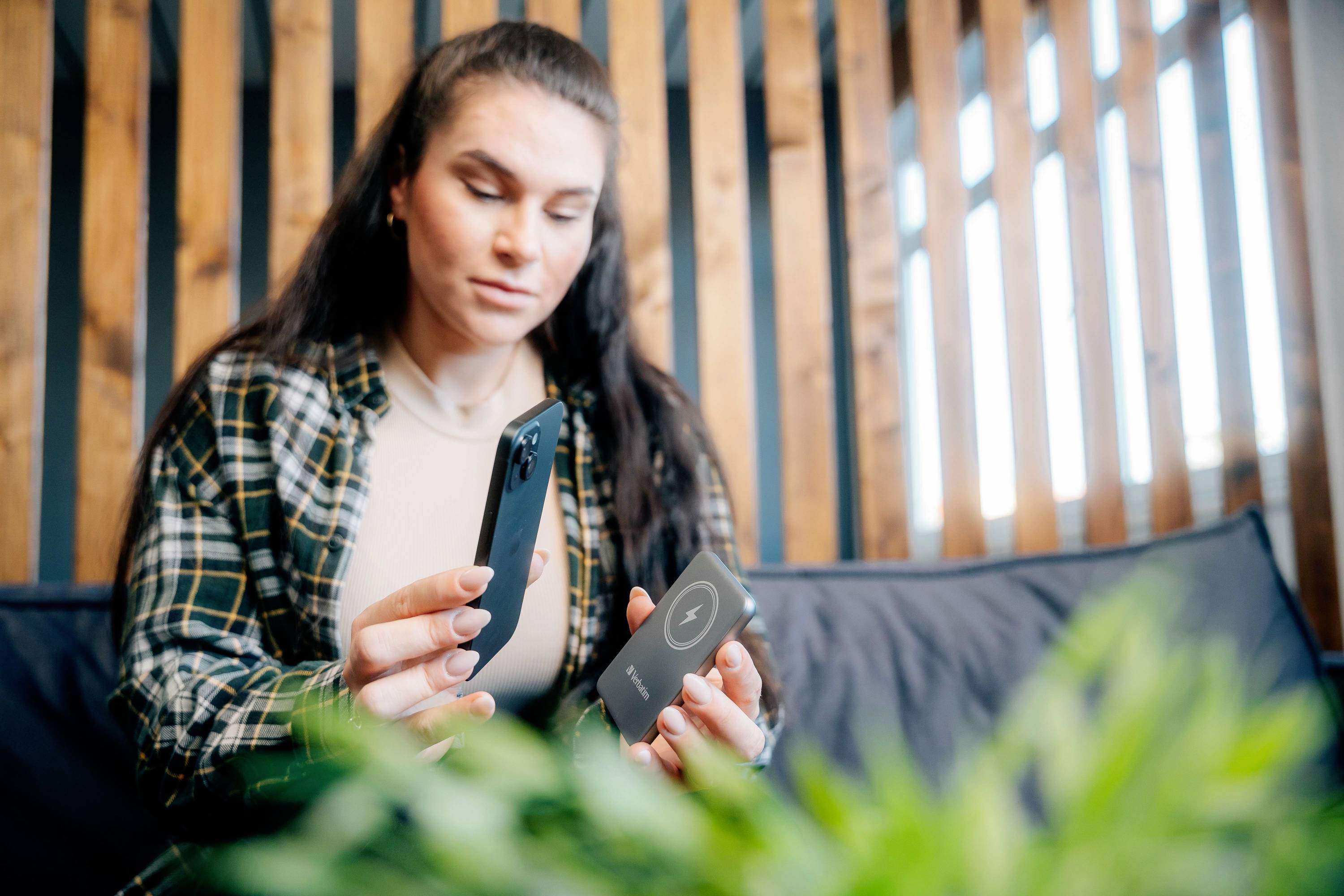 Une femme tient un smartphone et une batterie externe dans ses mains alors qu'elle est assise sur un canapé. Une plante est visible au premier plan.