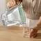 Une personne verse de l'eau d'une cruche dans un verre posé sur une table. L'image met l'accent sur l'action de verser.