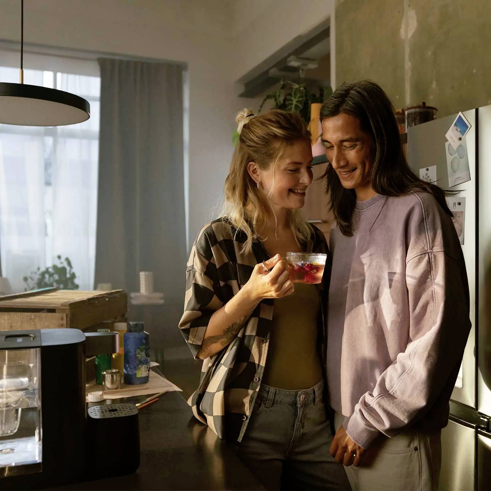 Une jeune femme et un jeune homme se tiennent souriants dans une cuisine, tandis que la femme tient une boisson. L'ambiance est chaleureuse et conviviale.