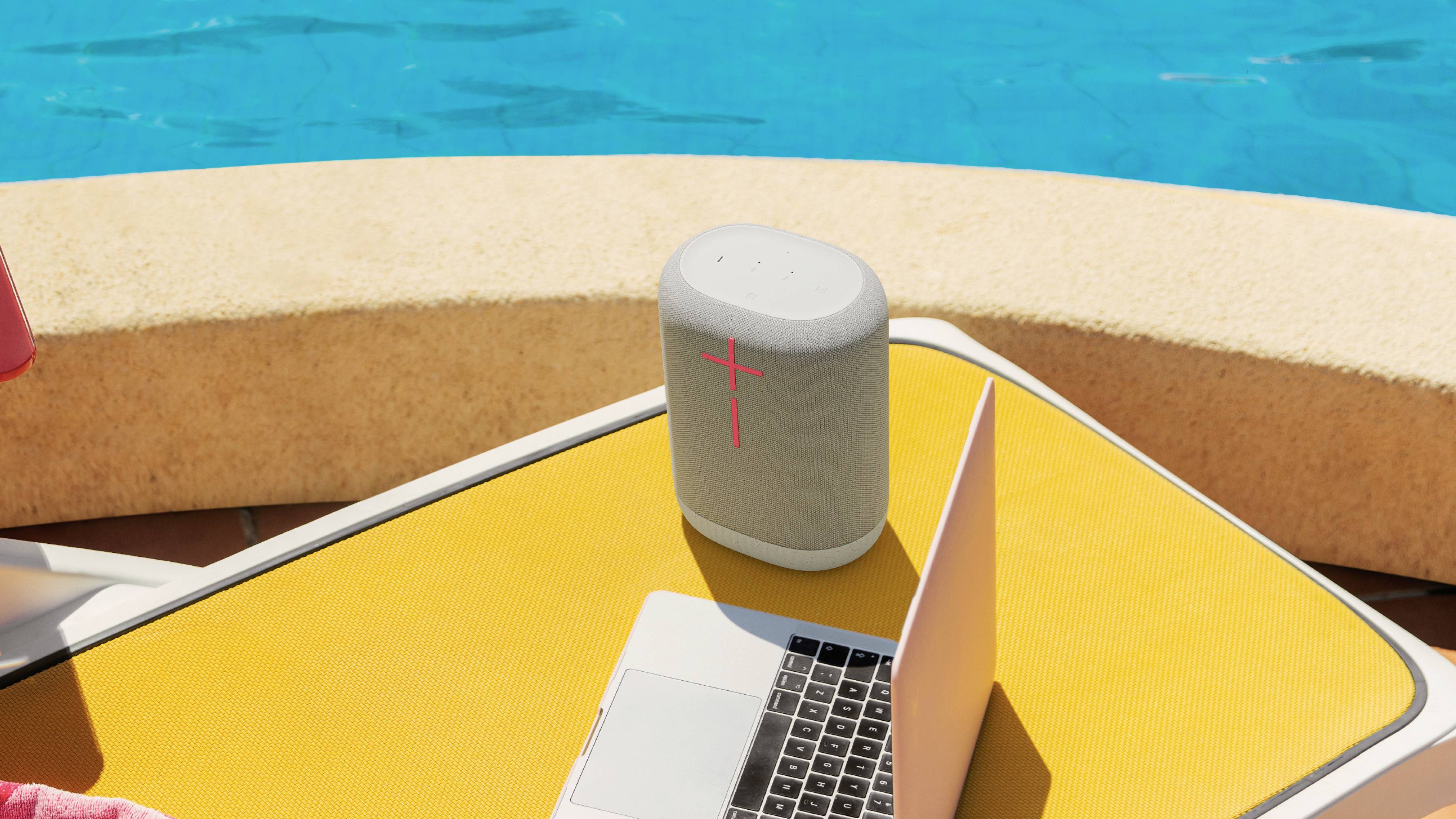 Un ordinateur portable et un haut-parleur sont posés sur un banc jaune à côté d'une piscine. Le haut-parleur affiche un signe plus rouge lumineux.