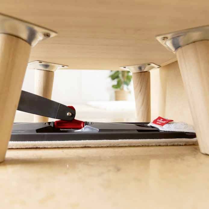 Un robot aspirateur aspire sous une table en bois. Un cache-pot est posé sur le sol en arrière-plan.