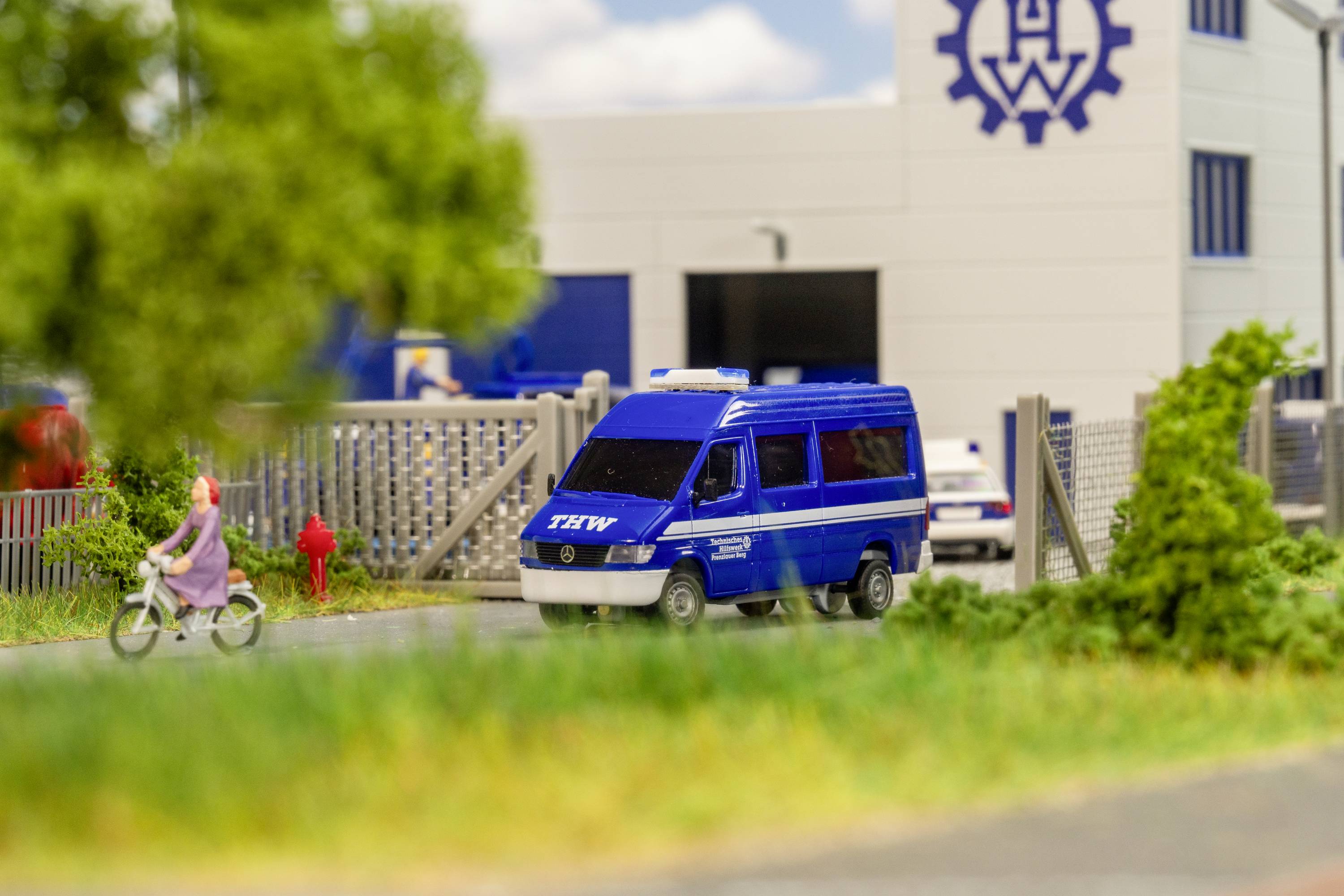 Une voiture d'intervention bleue du THW (Service technique de secours fédéral) est garée devant un bâtiment du THW. Une personne passe à vélo.