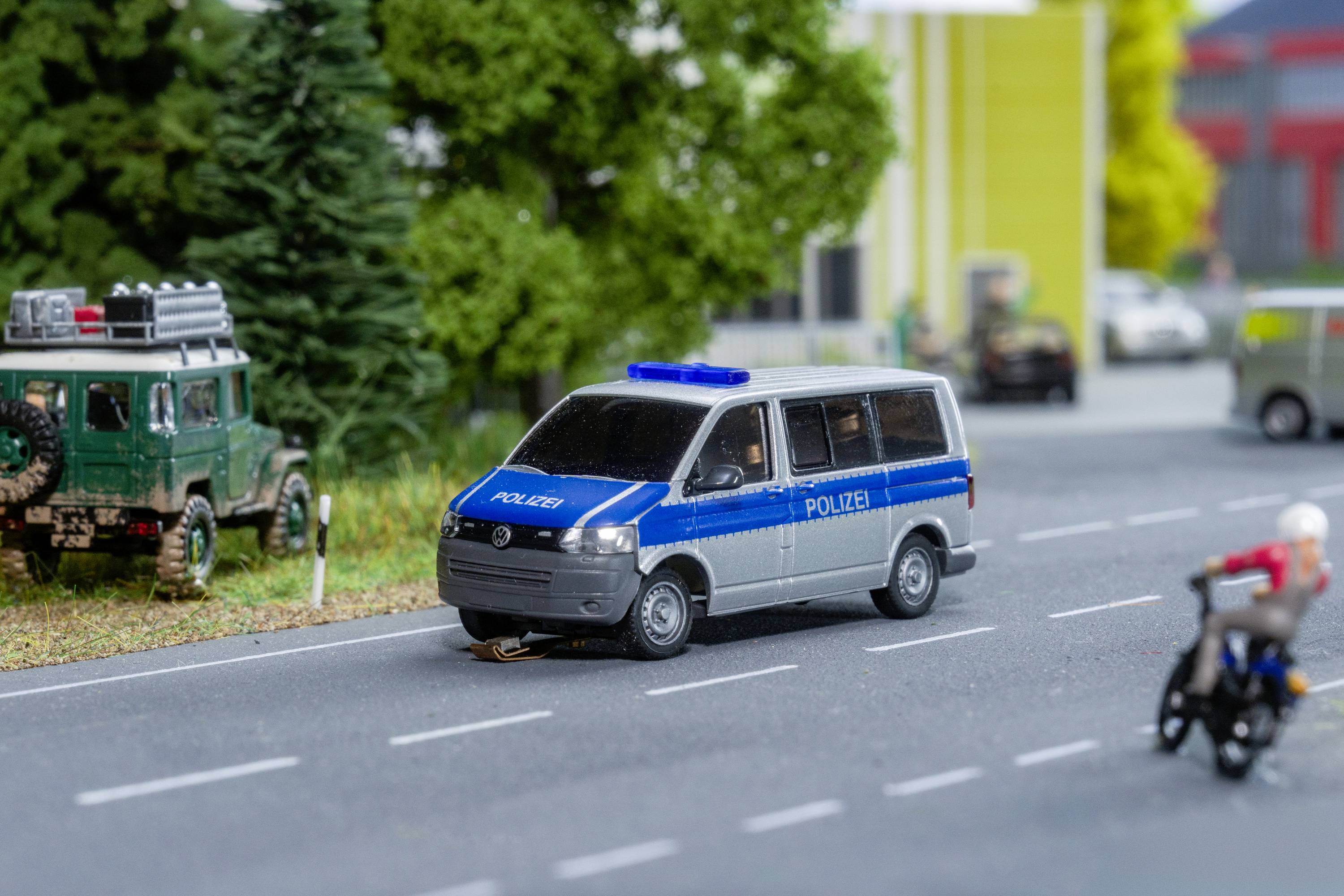 Un fourgon de police bleu et blanc est stationné sur une route de campagne. À l'arrière-plan, on aperçoit un tout-terrain vert et des bâtiments flous.