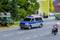 Un fourgon de police bleu et blanc est stationné sur une route de campagne. À l'arrière-plan, on aperçoit un tout-terrain vert et des bâtiments flous.