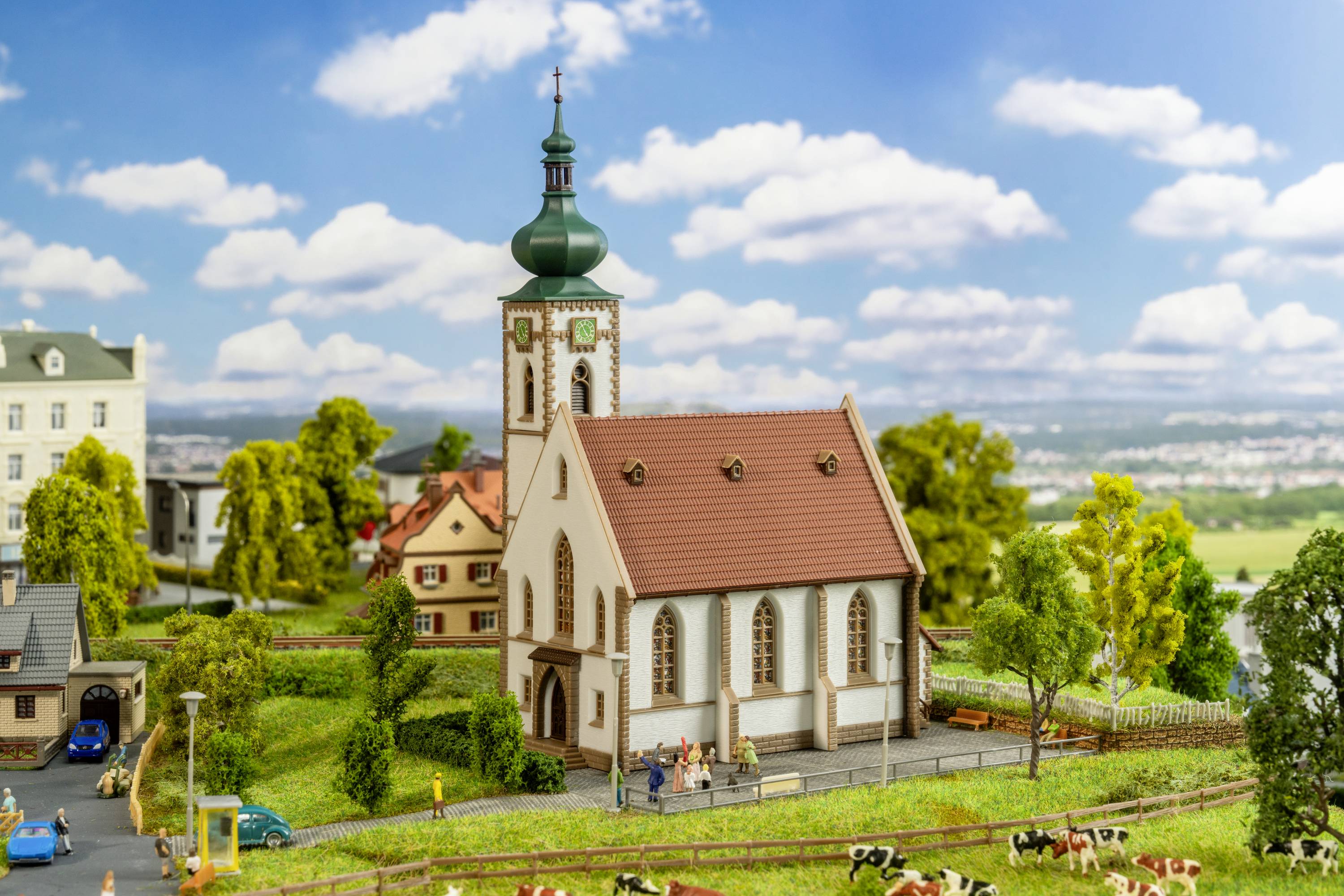 Un paysage miniature avec une église au centre, entourée de maisons et d'arbres, sous un ciel nuageux.
