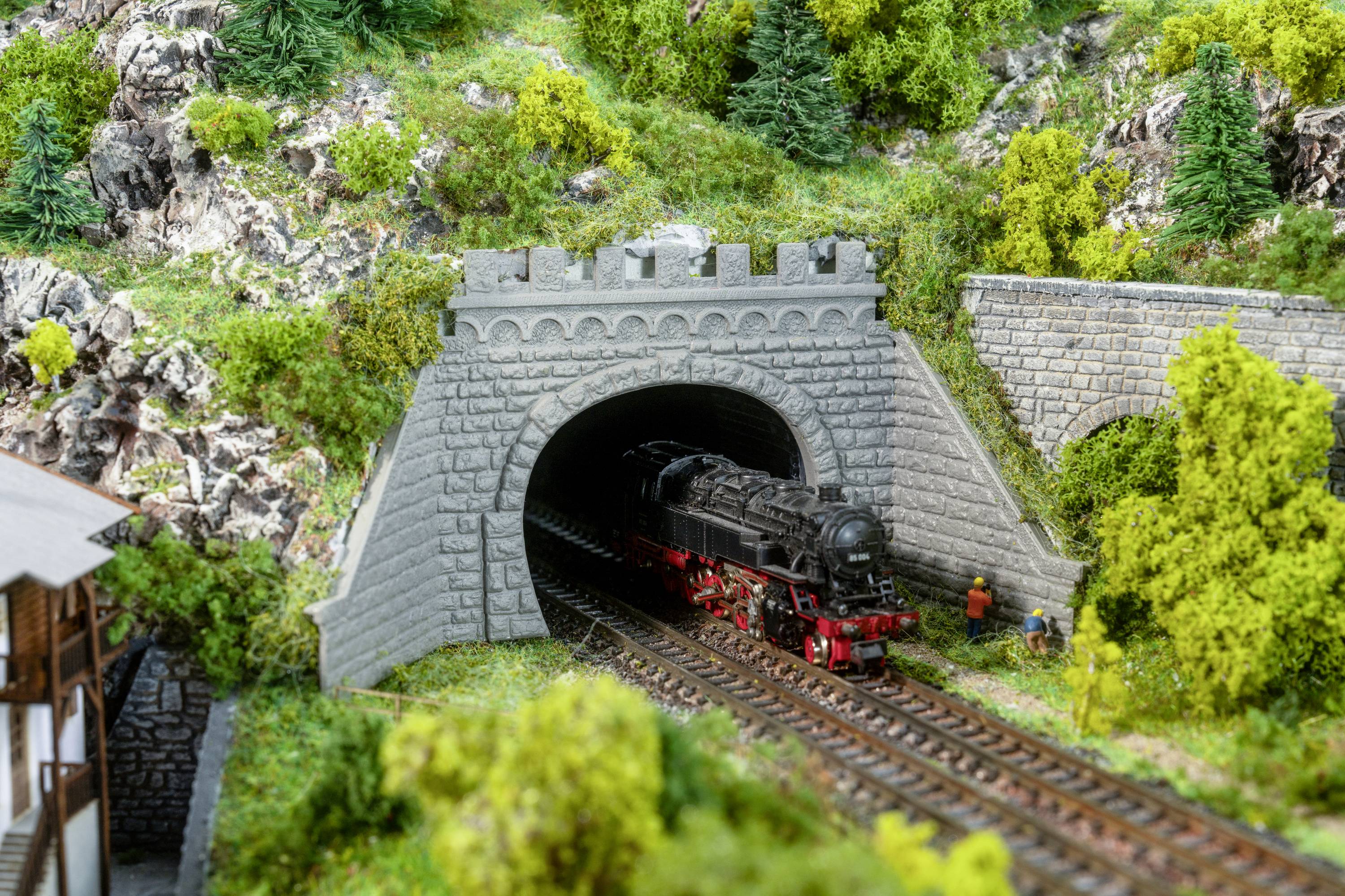 Un train miniature sort d'un tunnel dans un paysage montagneux. Au premier plan, deux personnages sont debout à côté des rails.