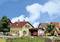 Un village idyllique aux maisons à colombages. Au premier plan, des personnes travaillant dans un jardin, une voiture rouge est garée dans l'allée.