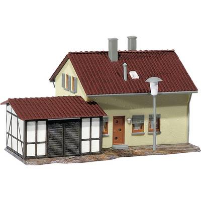Une maquette de maison avec un toit rouge et une façade jaune-brune, des bâtiments annexes avec un revêtement en bois, et un lampadaire devant.
