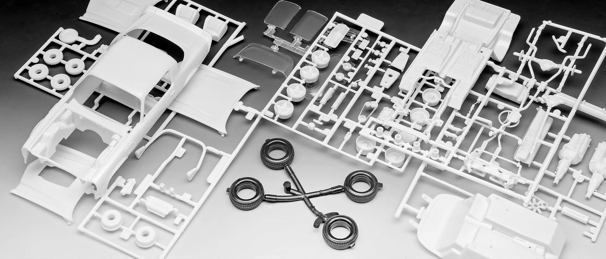 Kit de modélisme automobile composé de différentes pièces en plastique blanc et de pneus noirs sur une surface grise, prêt à être assemblé.