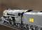 Un train miniature en métal aux détails soignés roule sur des rails. Le train est de couleur noire avec des autocollants jaunes portant des numéros.
