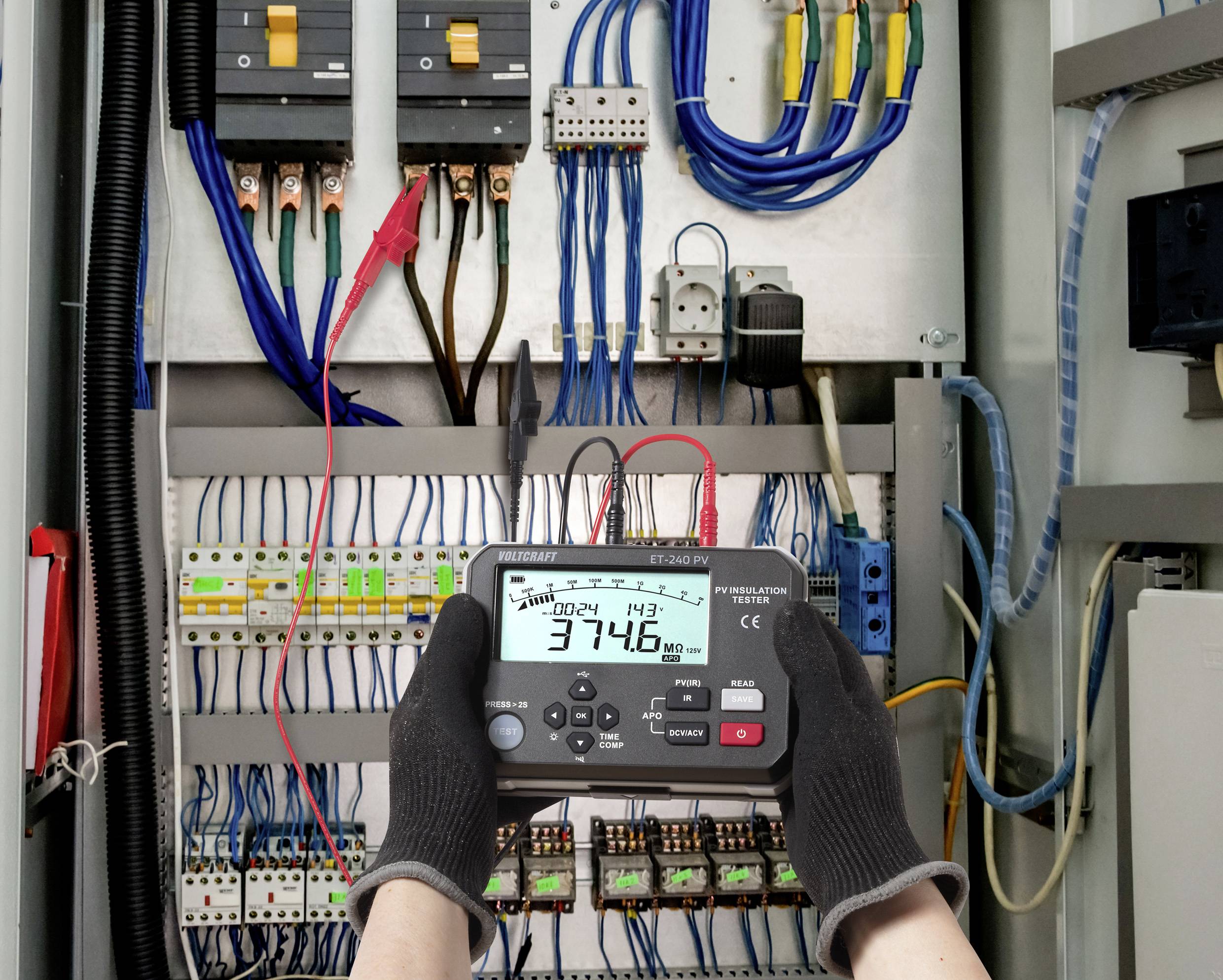 Personne utilisant un multimètre à l'intérieur d'un tableau électrique, mesurant 374,6 volts DC, entourée de fils, de disjoncteurs et de commandes.