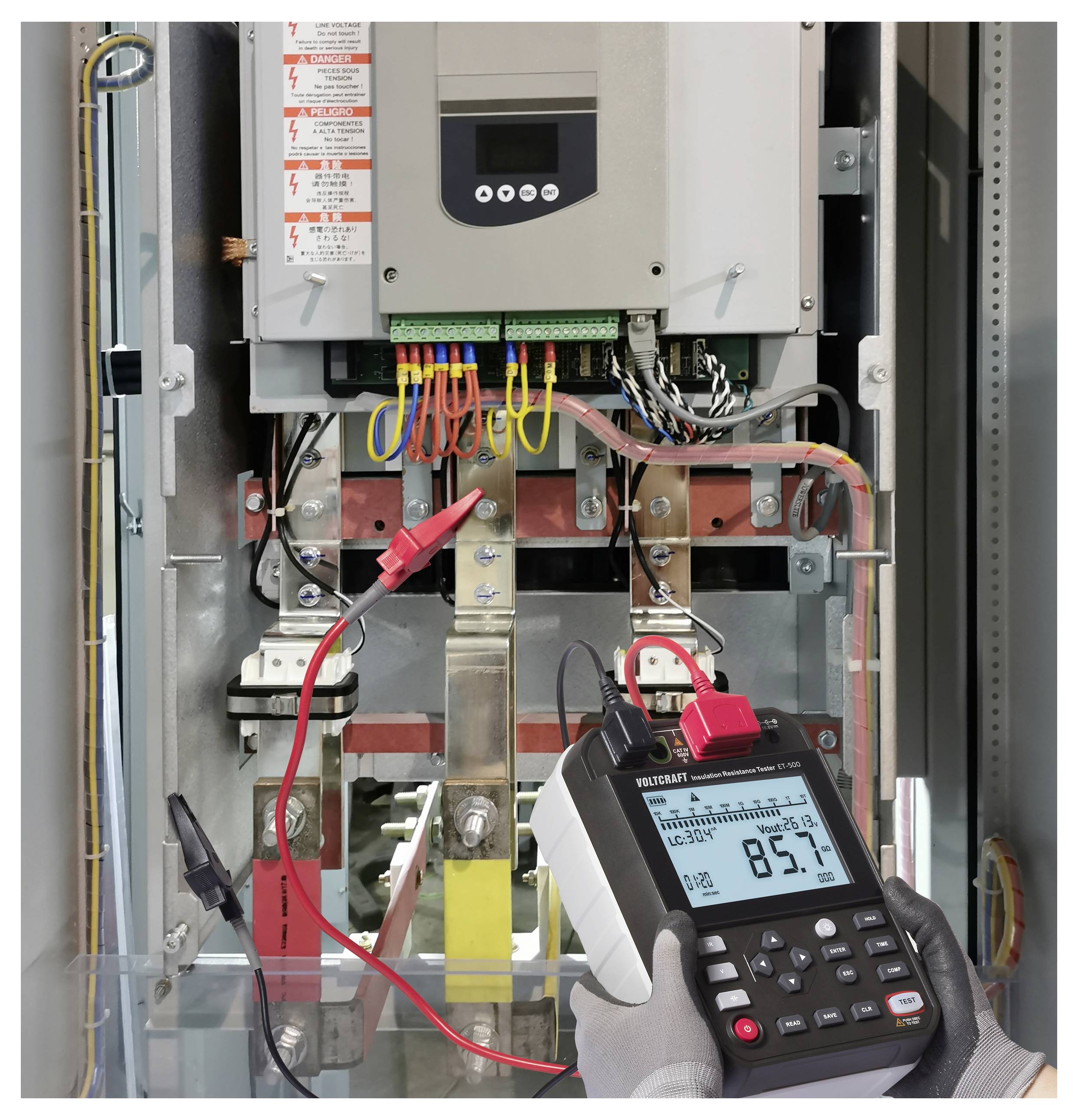 Une personne utilise un multimètre numérique pour mesurer la tension dans un panneau électrique, affichant une lecture de 85,7 volts. Des étiquettes de sécurité sont visibles.
