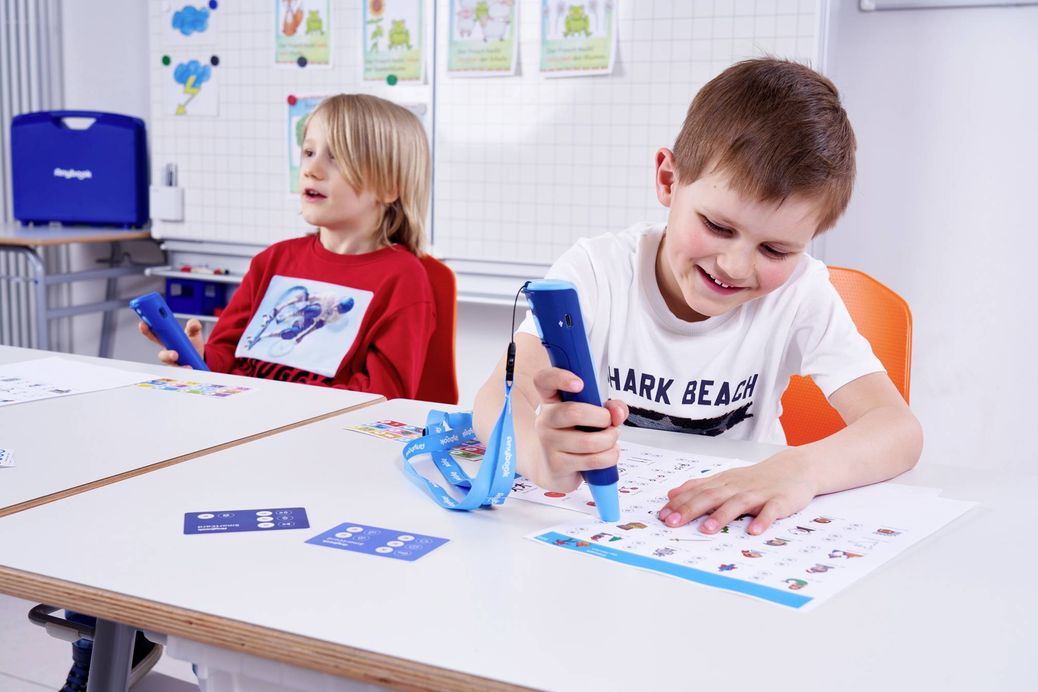 Deux enfants sont assis à des pupitres et utilisent des stylos éducatifs sur des feuilles de travail. Ils semblent concentrés et joyeux.