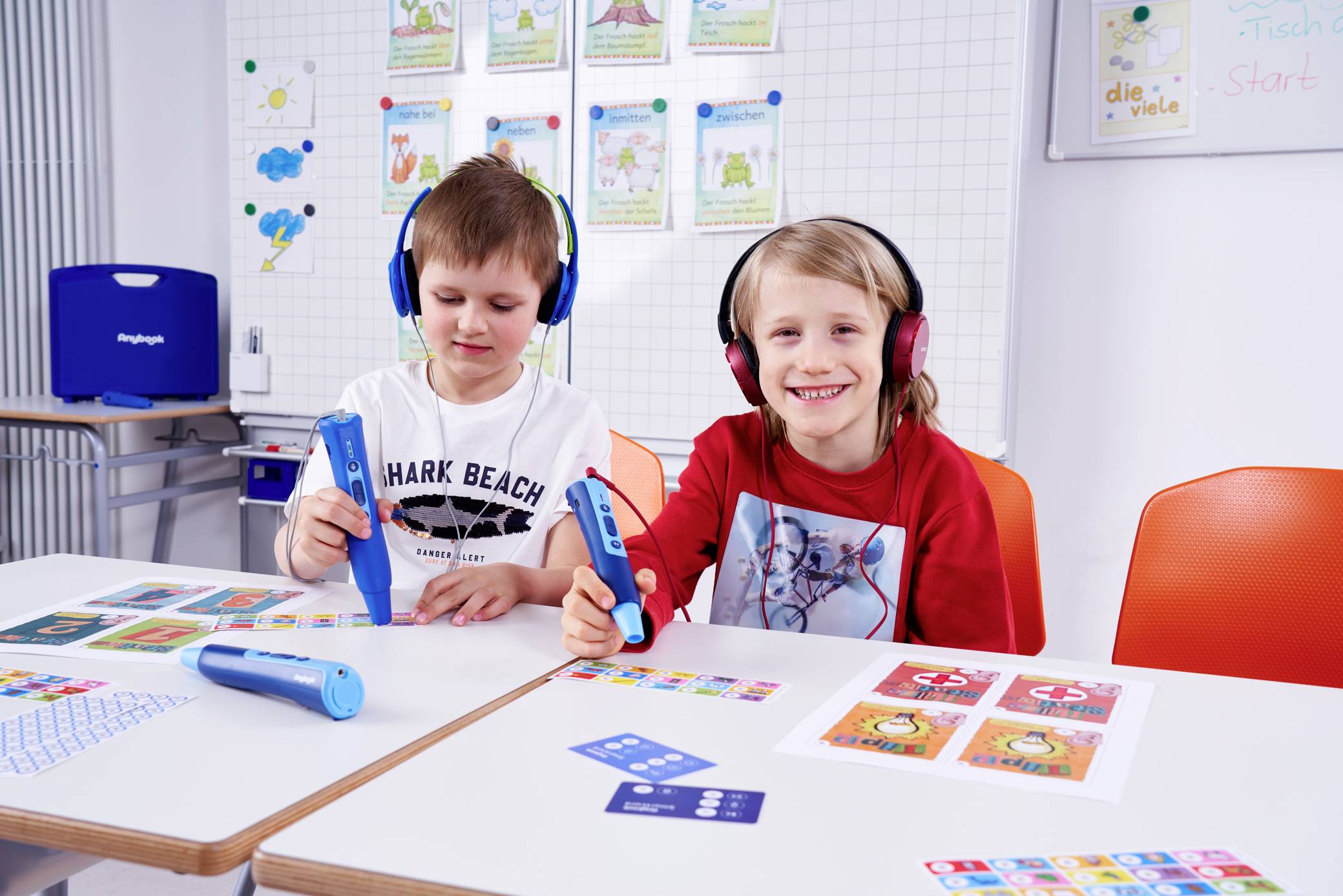 Deux enfants sont assis à une table et utilisent des stylos éducatifs. Ils portent tous deux des écouteurs. À l'arrière-plan, des affiches pédagogiques sont accrochées à un tableau.