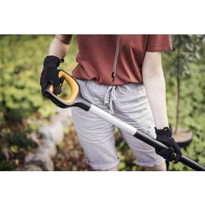 Une personne vêtue de vêtements de jardinage tient un outil de jardinage avec une poignée orange. Des forêts et des plantes sont visibles en arrière-plan.