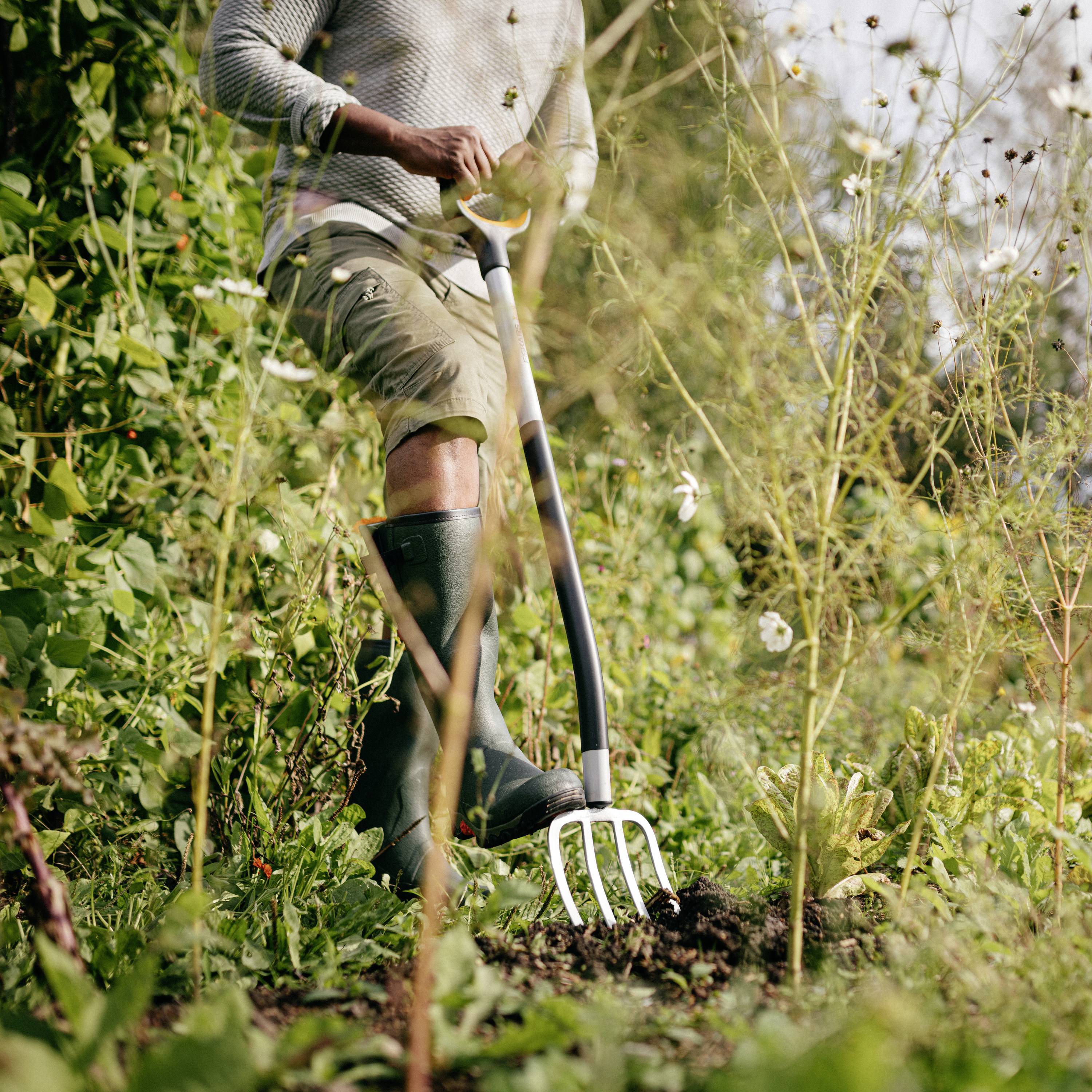 Une personne portant des bottes de caoutchouc retourne la terre dans un jardin, entourée de plantes vertes.