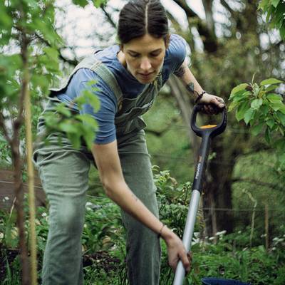 Une personne travaille dans le jardin. Elle porte des vêtements bleus et tient une pelle à la main. Des plantes et des arbres sont visibles en arrière-plan.