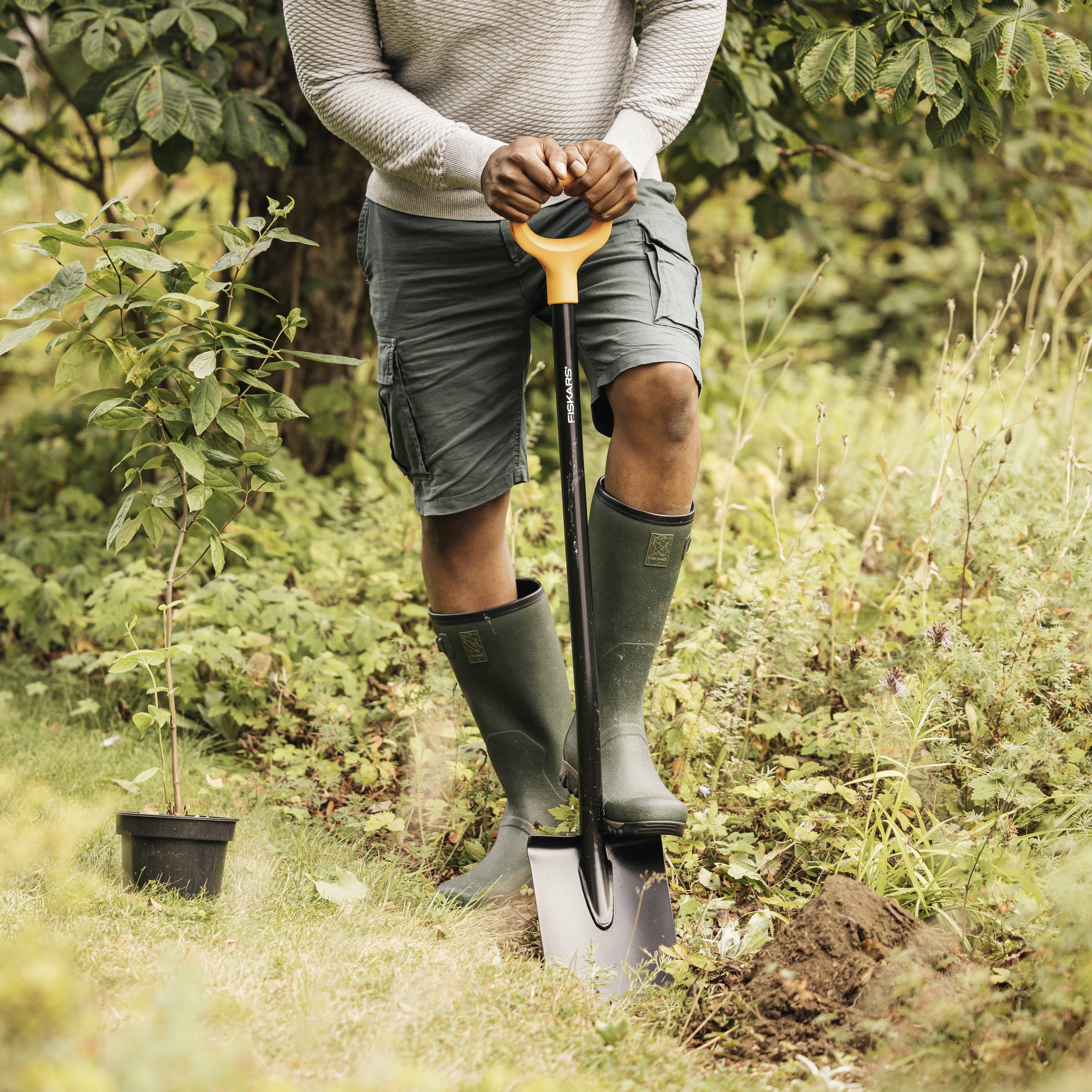 Un homme plante un petit arbre dans un jardin. Il porte des bottes en caoutchouc et tient une pelle. Autour de lui, il y a de la végétation herbeuse.