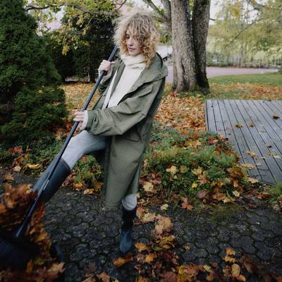 Une personne portant un manteau vert balaye des feuilles sur un trottoir en automne. En arrière-plan, on peut voir des arbres aux feuillages colorés.