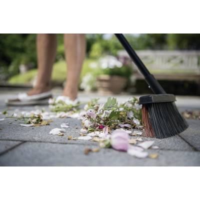 Une personne balaie des pétales et des feuilles d'une terrasse. À l'arrière-plan, on distingue des plantes de jardin floues et un banc en bois.