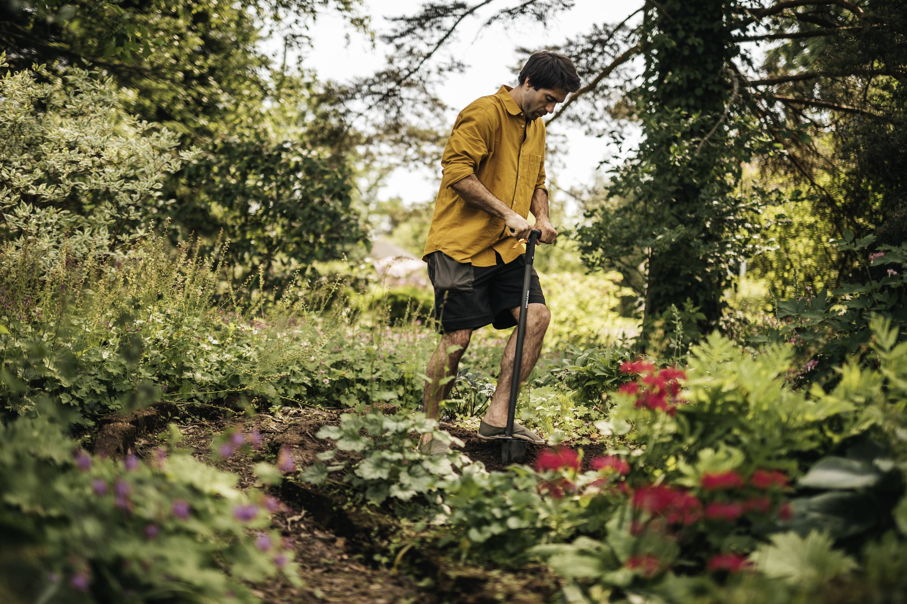 Une personne portant une veste jaune creuse avec une pelle dans un jardin vert et fleuri, avec des fleurs rouges et des arbres en arrière-plan.