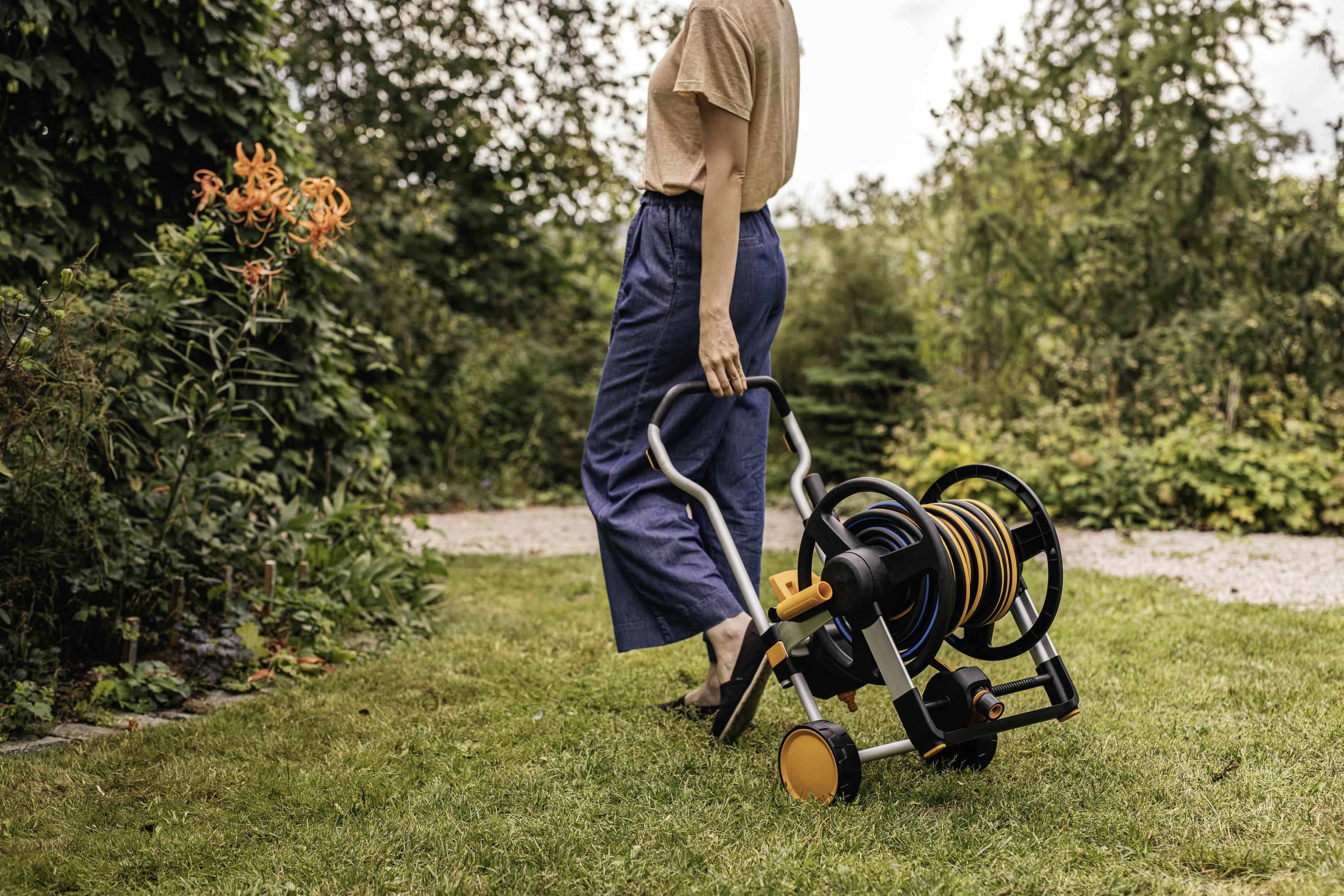 Une personne tire un tuyau d'arrosage sur une pelouse dans un jardin, entouré de buissons et de fleurs.