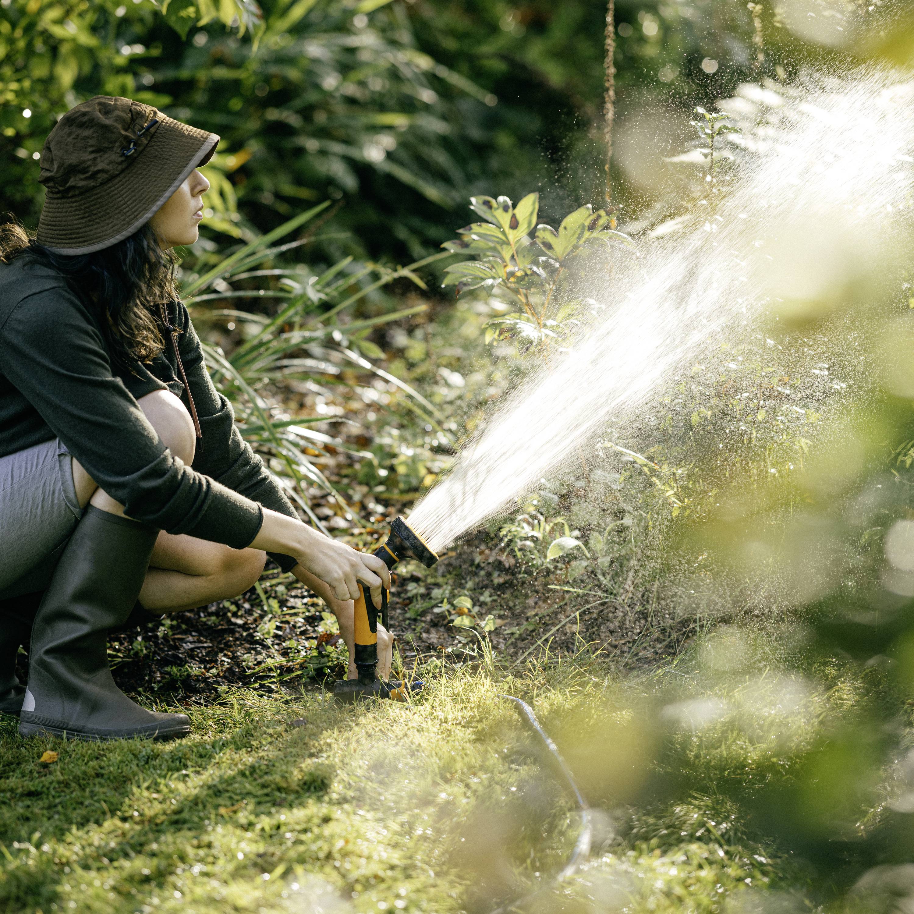 Une personne portant un chapeau de soleil arrose des plantes dans le jardin avec un tuyau d'arrosage. Entourée de végétation verdoyante et de lumière solaire.