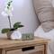 Une orchidée blanche dans un pot rond se trouve sur une table de chevet en bois à côté d'un réveil numérique qui affiche 13h45.