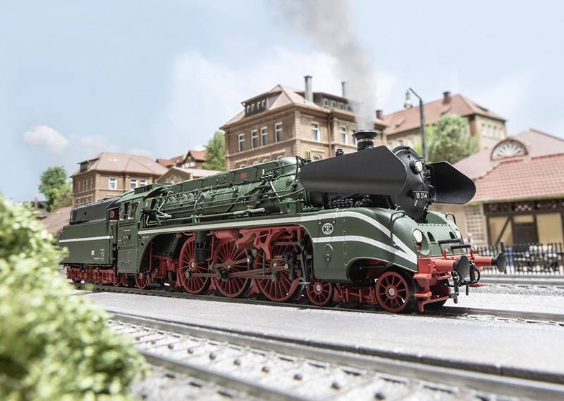 Un modèle vert d'une locomotive à vapeur historique circule sur des rails devant un arrière-plan avec des bâtiments et un ciel bleu.