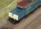Une locomotive miniature détaillée sur un rail, entourée d'herbe. La locomotive est peinte en bleu et crème.