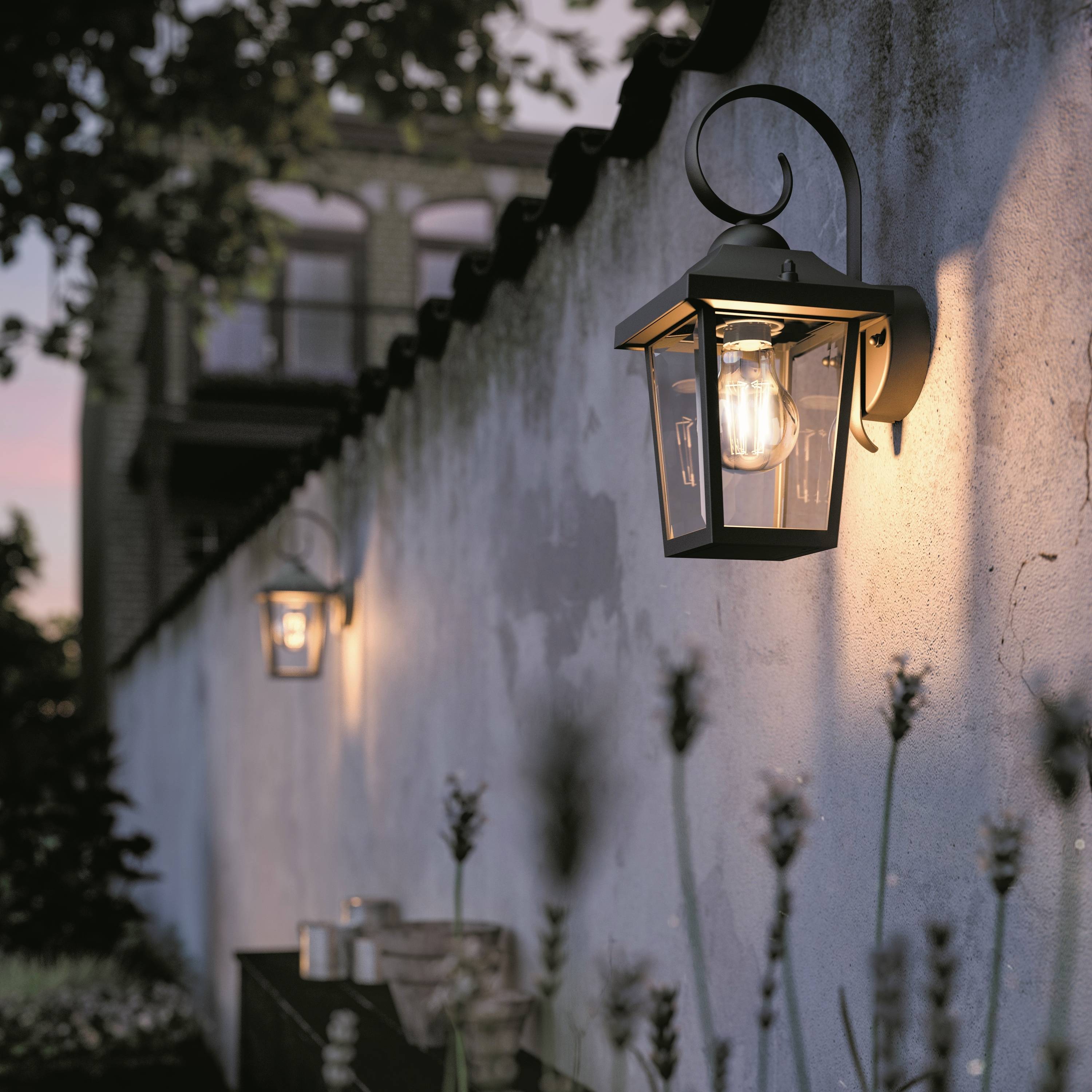 Des lanternes murales illuminent un mur de briques au crépuscule. Au premier plan, des plantes décoratives sont visibles.