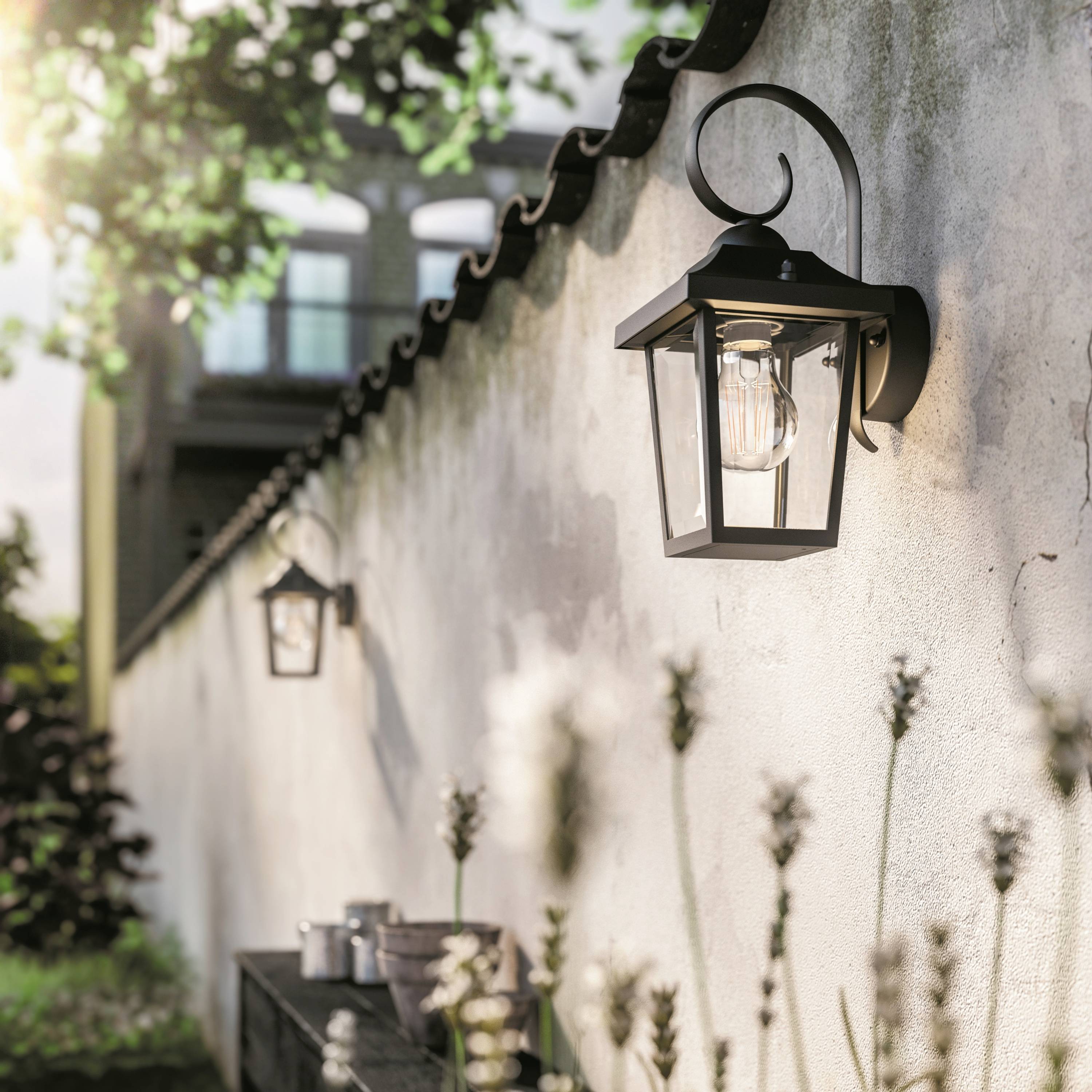 Un mur avec deux lanternes de style rustique qui brillent dans la lumière du soleil. Des plantes poussent au pied du mur, qui apparaît flou en arrière-plan.