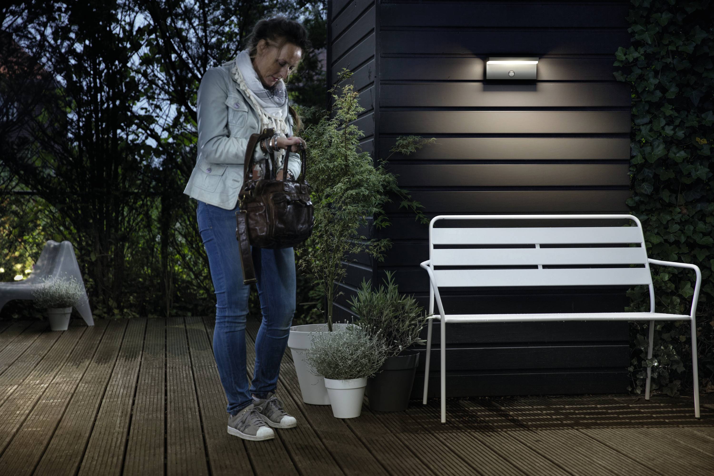 Une personne se tient debout la nuit sur une terrasse et regarde dans son sac. Un banc et des plantes sont visibles en arrière-plan.