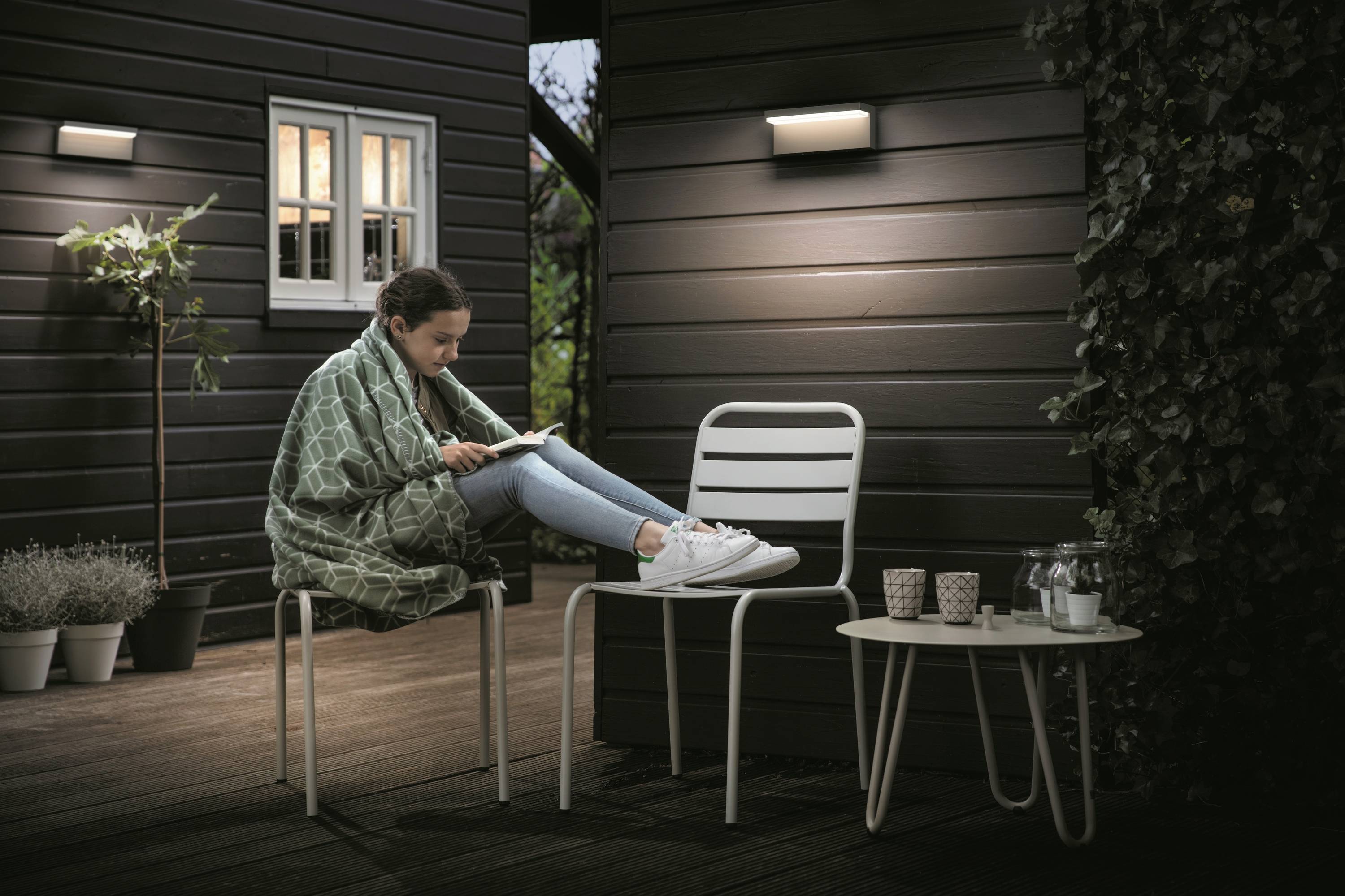 Une personne est assise dehors sur une chaise, enveloppée dans une couverture, en train de lire un livre. C'est le soir, éclairage au mur, plante à côté.