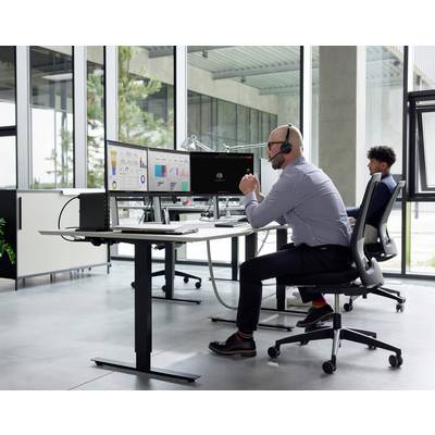 Un homme portant un casque de communication travaille à un bureau équipé de plusieurs écrans affichant des graphiques, tandis qu'une autre personne est assise à un bureau proche dans un bureau.