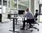 Un homme portant un casque de communication travaille à un bureau équipé de plusieurs écrans affichant des graphiques, tandis qu'une autre personne est assise à un bureau proche dans un bureau.