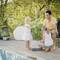 Un homme et deux enfants arrosent des plantes près d'une piscine. La scène dégage une atmosphère paisible et estivale en plein air.