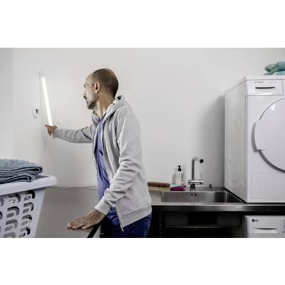 Un homme dans une buanderie allume une lampe murale lumineuse pendant qu'il se tient à côté d'une machine à laver. Dans la pièce se trouvent un lavabo et un panier à linge.