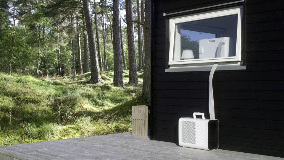 Un climatiseur portable se trouve sur une terrasse en bois devant une maison noire avec un arrière-plan boisé. Un tuyau mène vers la fenêtre.