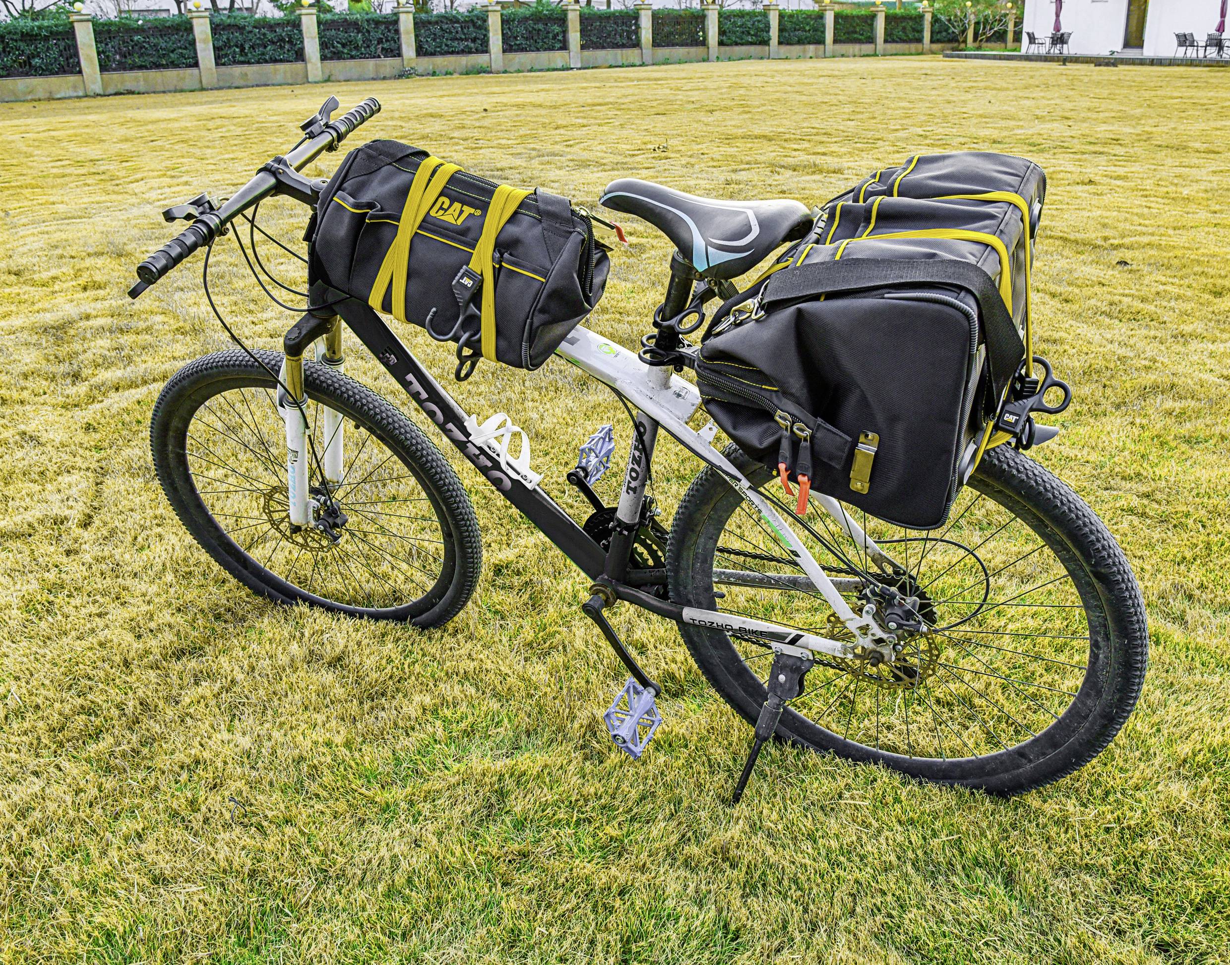Un vélo équipé de grandes sacoches noires et d'un sacoche de guidon, stationné sur de l'herbe sèche dans un parc avec une clôture en arrière-plan.