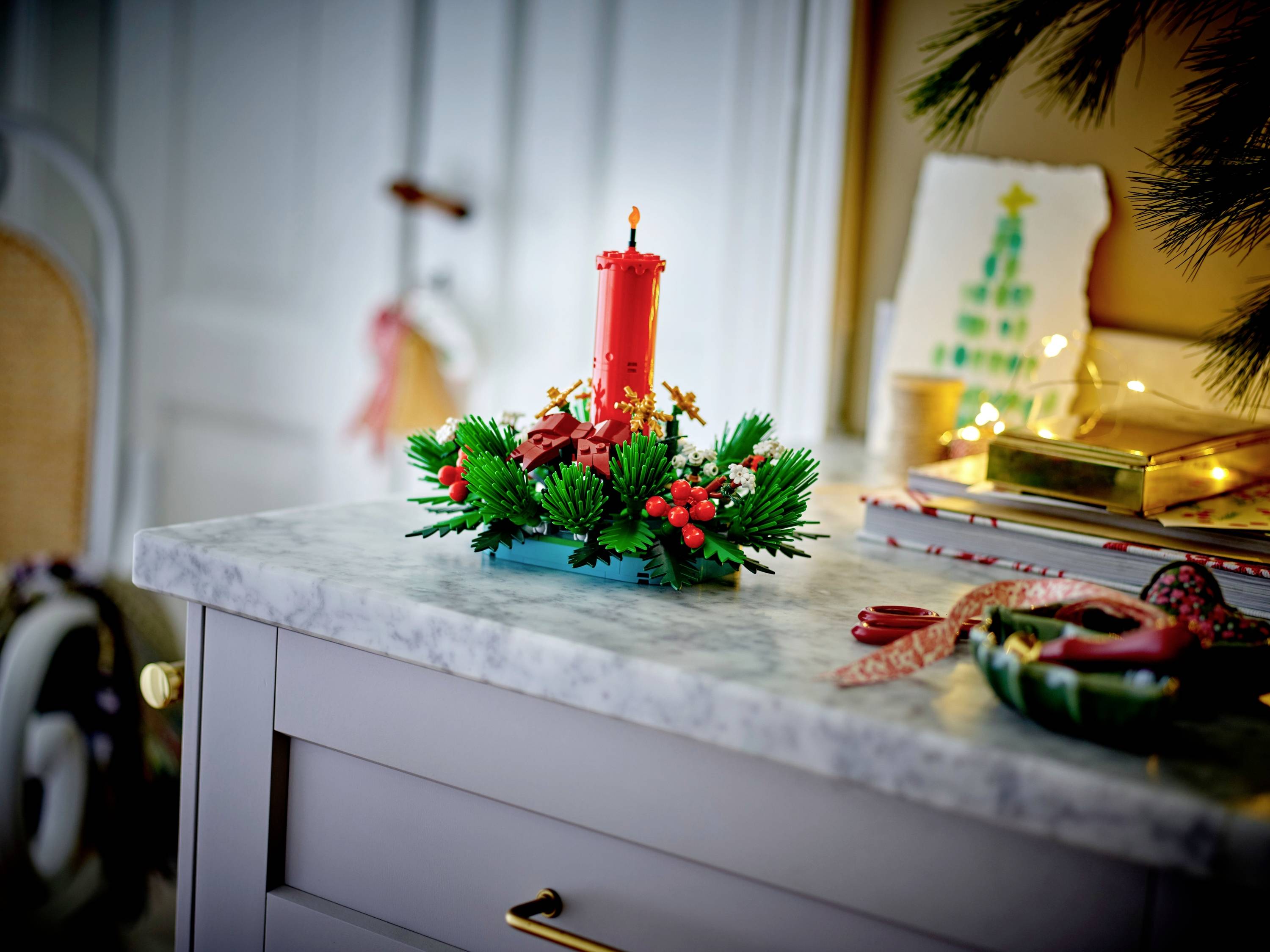 Une bougie festive décorée avec des baies rouges et du feuillage vert est posée sur un comptoir en marbre, à côté d'une carte représentant un arbre de Noël peint.