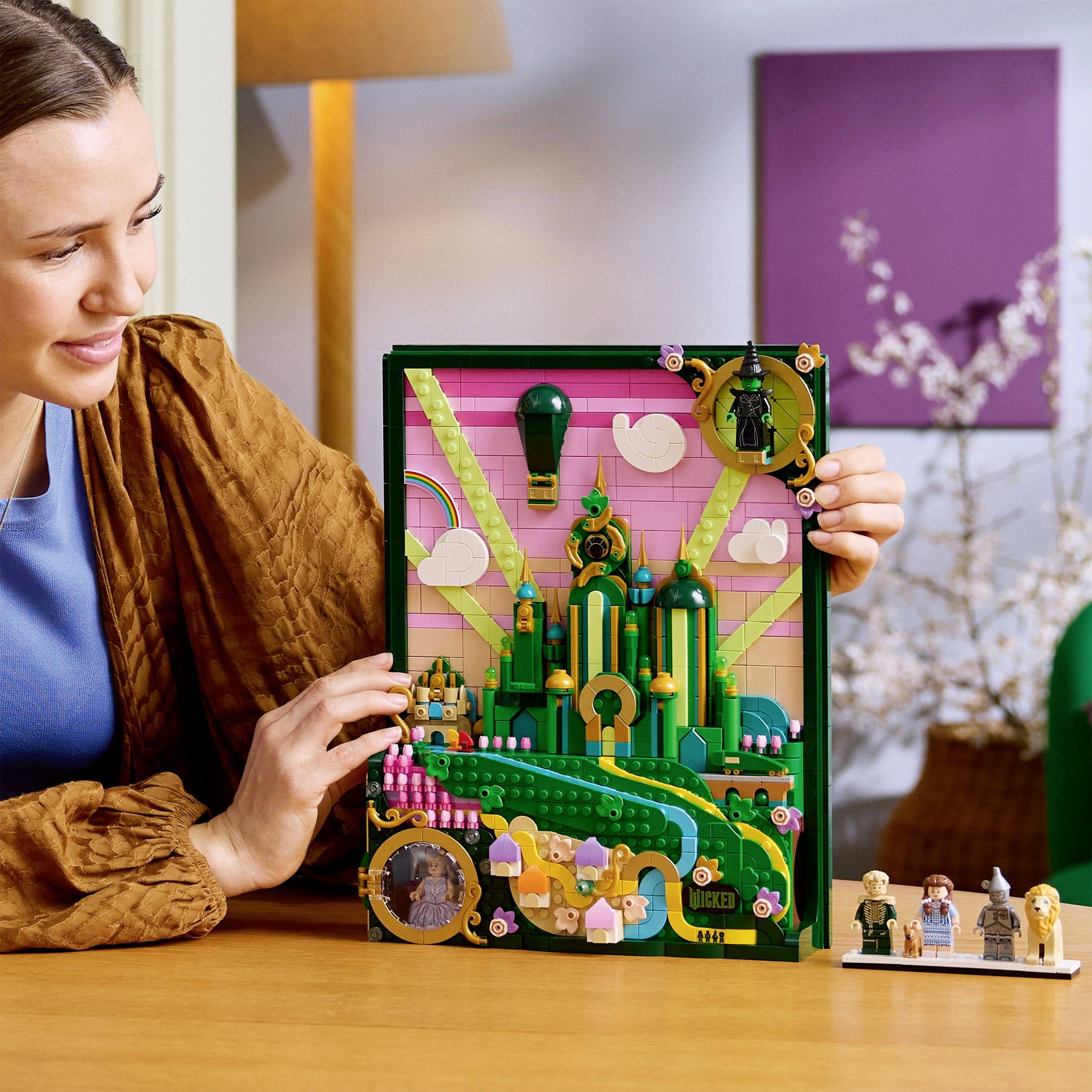Une personne sourit devant un ensemble de blocs de construction colorés qui évoque un paysage, avec de petits personnages disposés à la base.