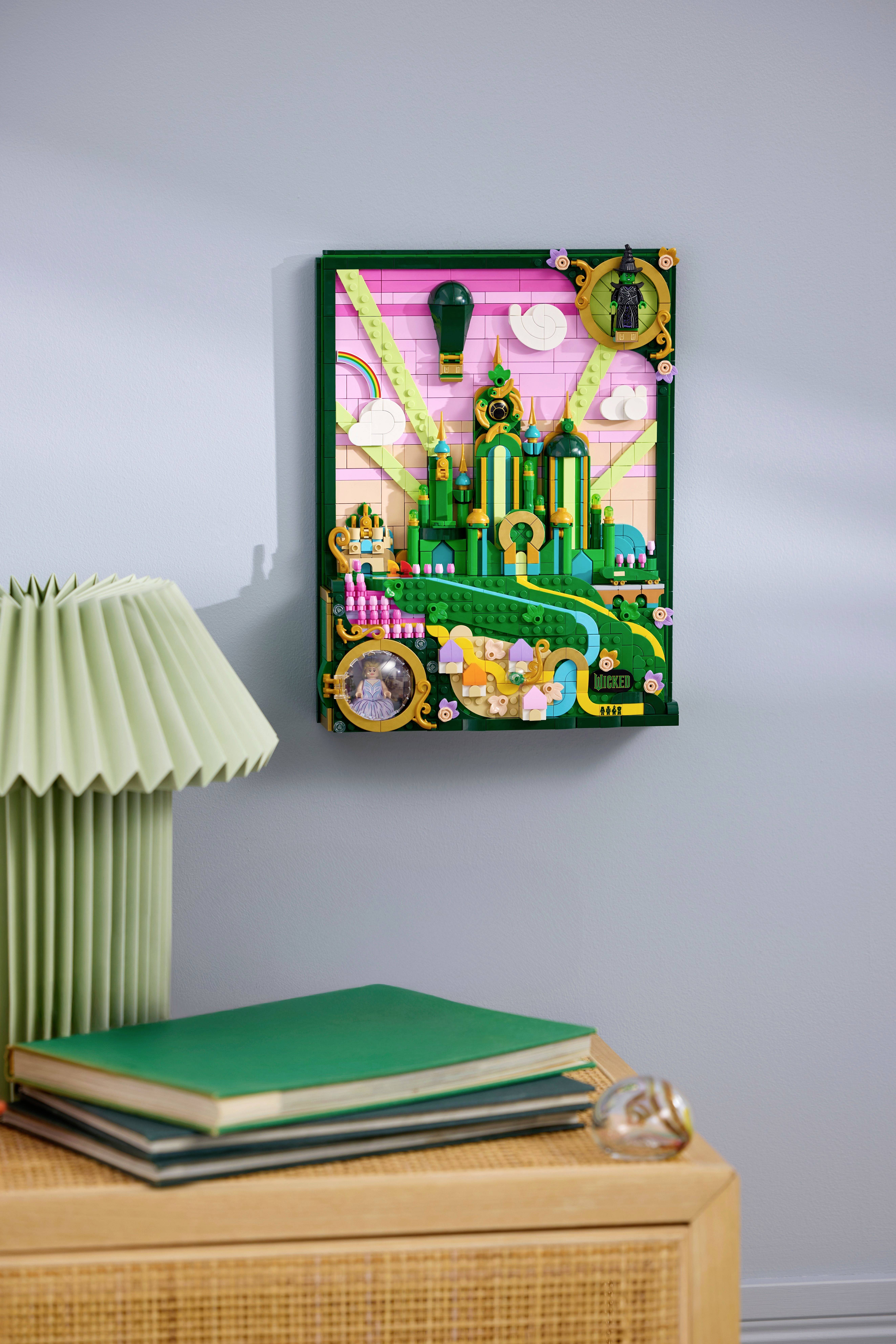 Une œuvre d'art colorée représentant un paysage urbain fantaisiste avec des tours et des chemins, encadrée de vert, est suspendue au mur au-dessus d'un bureau chargé de livres.