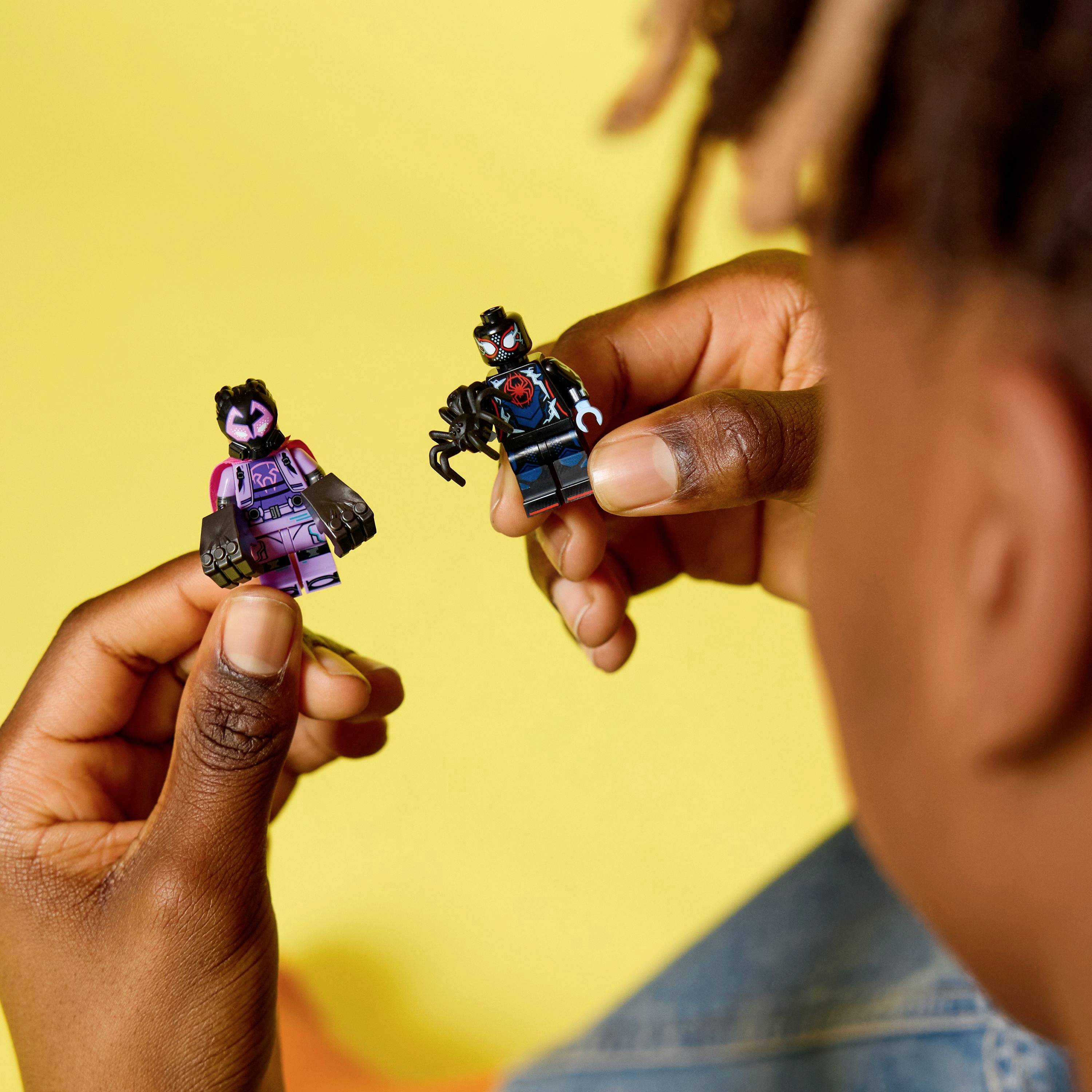 Une personne tenant deux figurines sur le thème des superhéros, une dans chaque main, devant un fond jaune vif.