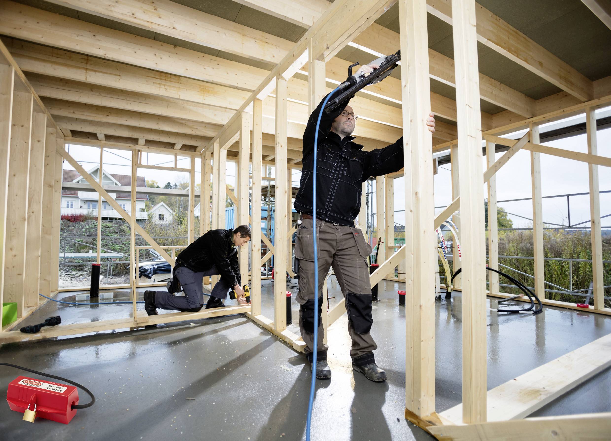 Deux ouvriers du bâtiment encadrent un mur en bois à l'intérieur d'une maison en cours de construction. L'un est debout avec un outil, l'autre est agenouillé, alignant des poutres.