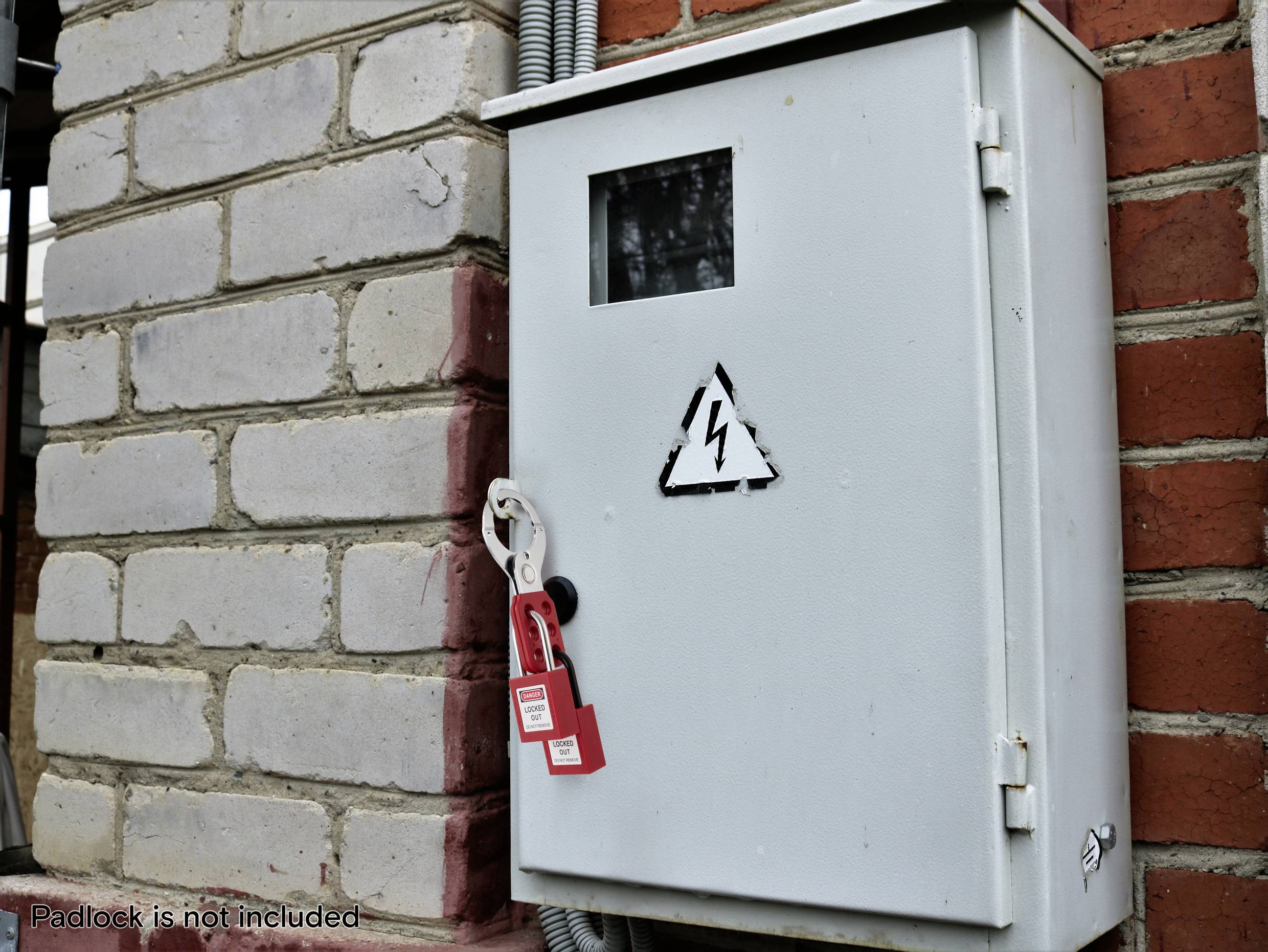 Boîte électrique en métal avec un symbole de foudre sur un mur de briques, scellée avec une étiquette de condamnation rouge. Texte d'avertissement : Cadenas non inclus.