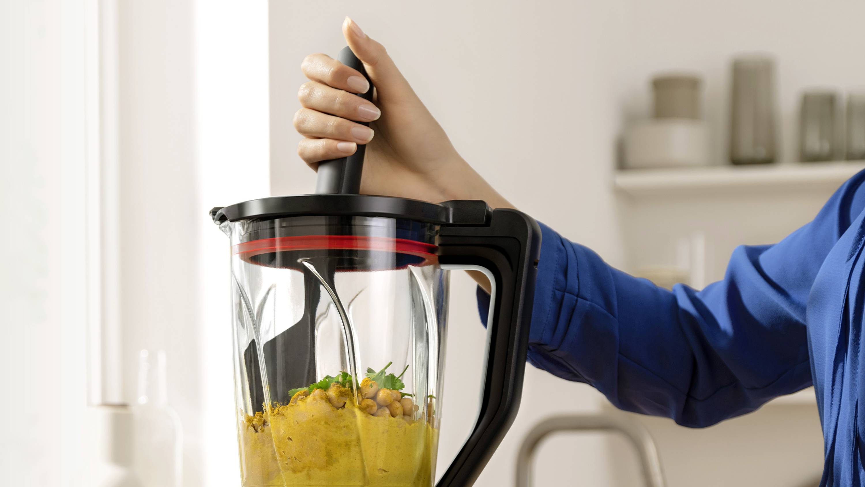 Une personne portant une chemise bleue utilise un mixeur à manche noir pour mélanger des pois chiches et des herbes, préparant une purée lisse dans un environnement de cuisine.